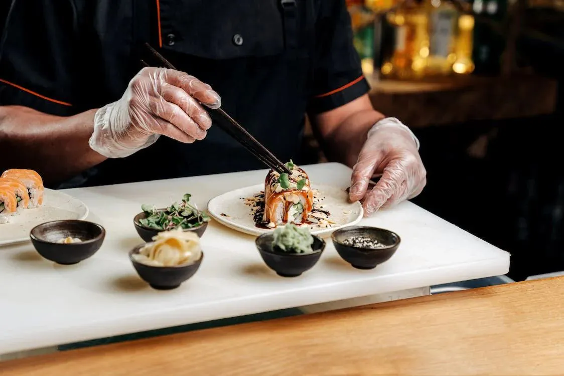 Sushi chef wearing gloves garnishes prepared sushi dish with chopsticks